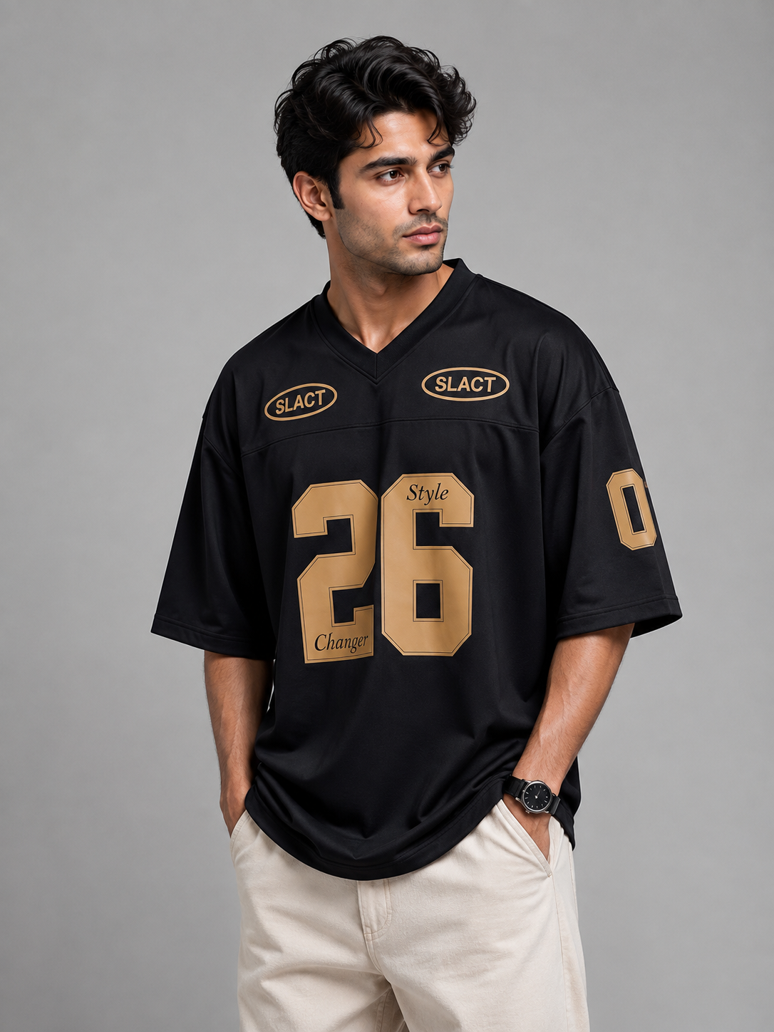 A man wearing a black athletic-style jersey with gold lettering and numbers, including the number 26 on the front and 'SLACT' logos on the chest, paired with light beige pants, against a solid gray studio background.