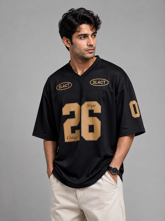 A man wearing a black athletic-style jersey with gold lettering and numbers, including the number 26 on the front and 'SLACT' logos on the chest, paired with light beige pants, against a solid gray studio background.