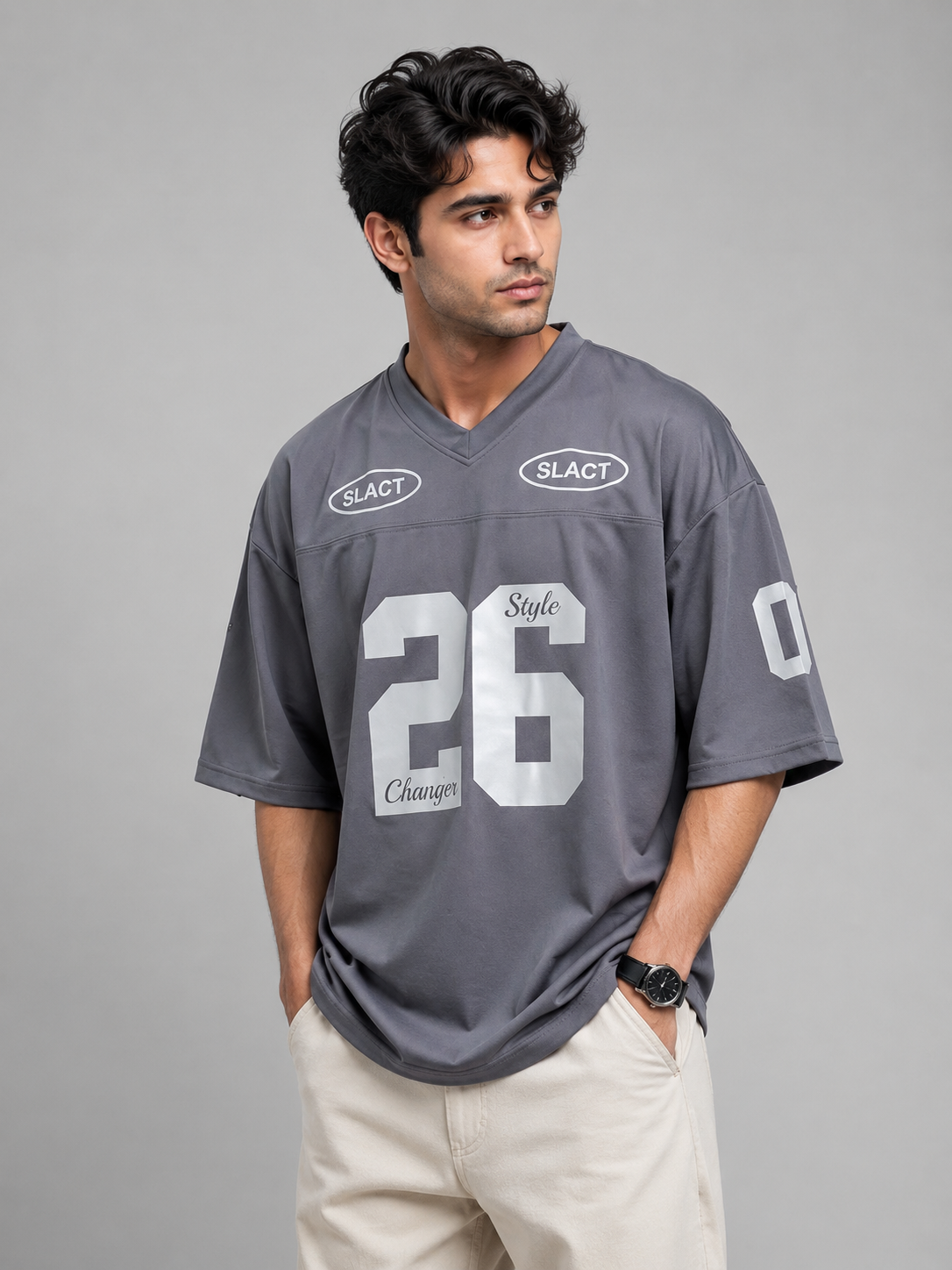 A man wearing a gray athletic-style jersey with white numbers and logos, paired with beige pants, standing against a solid gray studio background.
