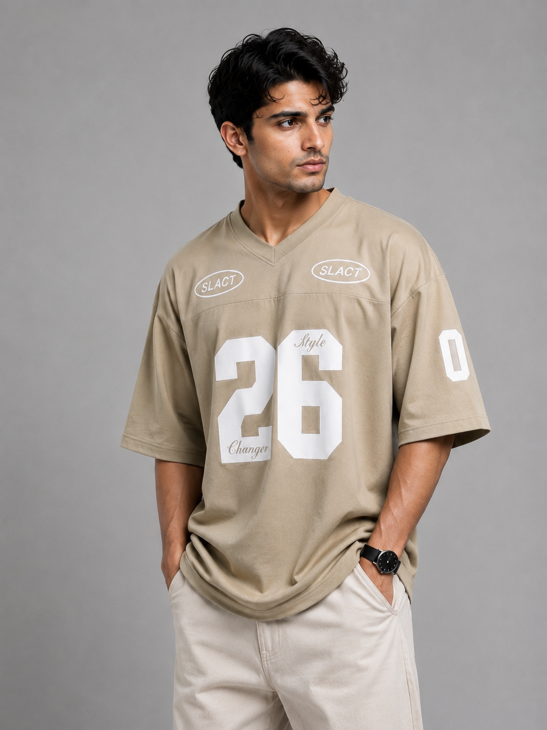A man wearing a beige v-neck athletic jersey with white numbers and logos, paired with beige pants, standing against a solid gray studio background.